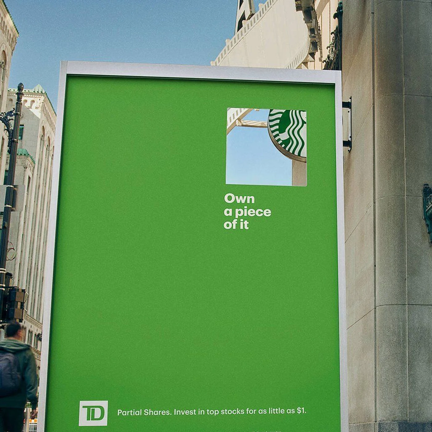 A green TD Bank billboard with a square window cut through it, framing part of the Starbucks siren on the building behind.